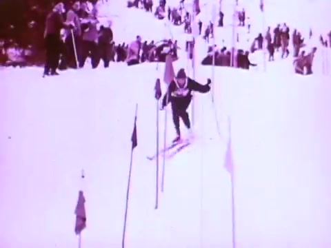 The still from the Mobil I oil commercial shows a skier navigating through a slalom course marked by flags on a snowy slope. A crowd of spectators is visible in the background, creating a lively atmosphere. The scene emphasizes action and excitement, likely highlighting the performance and reliability of Mobil I oil in extreme conditions. The image has a vintage quality, typical of 16mm film.