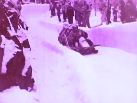 The still features a snowy landscape where a sledding team races down a narrow, icy track surrounded by spectators. The image has a distinct purple tint, emphasizing the cold, wintery setting. The focus is on the excitement of the competition, showcasing the performance of the sled, likely highlighting the importance of Mobil I oil in ensuring optimal vehicle performance in extreme conditions.