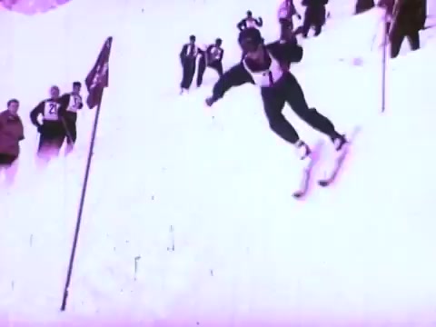 The still shows a skier mid-action, speedily descending a snowy slope. The skier is dressed in a black and white outfit and appears focused on navigating a ski race marked by flags. In the background, several spectators watch the event. The overall atmosphere conveys excitement and energy, highlighting the performance of the skier against a winter backdrop, consistent with the themes of precision and quality often associated with Mobil I oil commercials.