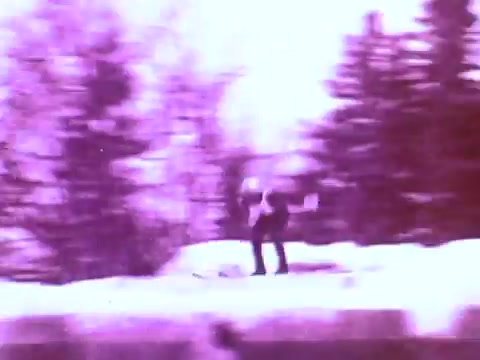 The still from the Mobil I oil commercial captures a dynamic scene of a skier in mid-motion, skiing along a snowy landscape. The image has a distinctive purple tint, giving it a vintage aesthetic. Snow-covered trees provide a backdrop, emphasizing a winter environment. The skier appears to be performing an athletic maneuver, showcasing speed and agility, typical of sports commercials that highlight performance and durability, aligning with the brand's focus on high-quality oil.