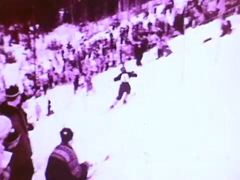 The still shows a skier racing down a snowy slope, with a crowd of spectators cheering on the sides. The image has a vintage look, typical of 16mm film, featuring a pinkish hue. The scene captures the excitement of winter sports, likely emphasizing performance and adventure, aligning with the Mobil I oil commercial's theme of quality and reliability.