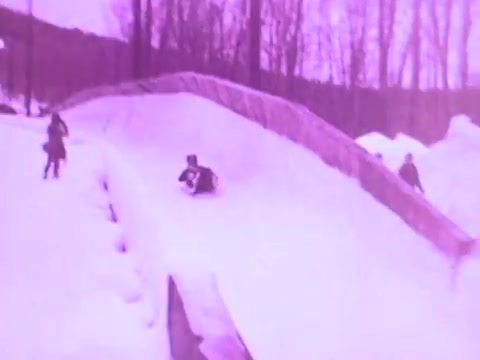 The still depicts a snowy landscape with people enjoying winter activities, possibly sledding. The predominant pink tint gives it a vintage look, characteristic of 16mm film. The setting emphasizes a playful and adventurous feel, aligning with themes of durability and performance often highlighted in oil commercials.