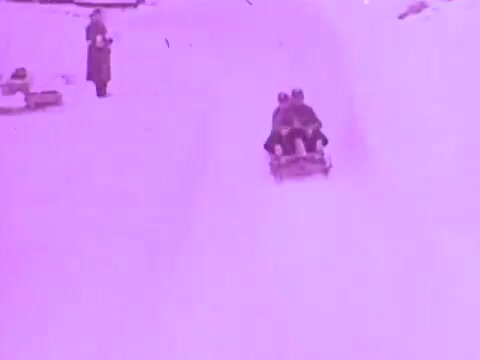 The still depicts a snowy landscape, capturing a lighthearted moment where two individuals are sledding down a snow-covered hill. A person in the background appears to be observing the action, while the overall scene conveys a sense of winter fun and leisure, fitting the theme of the Mobil I oil commercial, which likely emphasizes performance and reliability in cold conditions. The color tone has a pinkish hue.