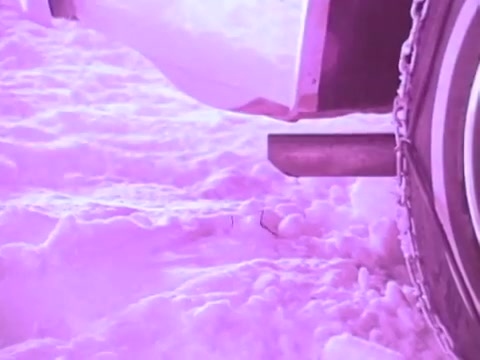 The still from the Mobil I oil commercial features a close-up view of a vehicle's exhaust pipe, with snow surrounding the tires. The scene has a pinkish tint, suggesting a cold environment, which emphasizes the product's performance in extreme conditions. This visual likely aims to highlight the oil's ability to maintain engine efficiency during winter drives.