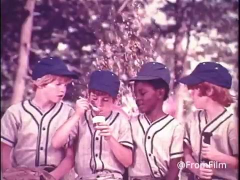 The 16mm still from the 1970s Hunt's Snack Pack Chocolate Pudding commercial features four boys in baseball uniforms, sitting together outdoors. They are wearing matching blue caps and appear to be enjoying their treats. One boy is holding a Snack Pack container and using a spoon to scoop out the pudding. The scene exudes a playful, nostalgic vibe typical of children's outdoor activities during that era, highlighting the fun and enjoyment associated with Snack Pack pudding. The tagline emphasizes making pudding a party, reflecting the brand's marketing approach.