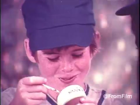 The still features a boy in a baseball cap, smiling as he enjoys a bowl of Hunt's Snack Pack Chocolate Pudding. His expression conveys delight, emphasizing the fun associated with the product. The backdrop gives a nostalgic feel, typical of advertisements from the 1970s. This captures the playful vibe of childhood and the excitement of enjoying snack foods, resonating with the theme of innocence prevalent in the era depicted, similar to the aesthetics in 'Stranger Things.'
