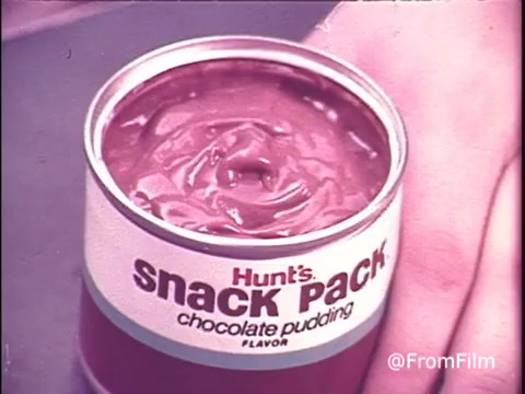 The 16mm still features a classic aluminum can of Hunt's Snack Pack Chocolate Pudding, typical of the 1970s. The colorful branding highlights the product's appeal, emphasizing 'snack pack' in bold letters. The pudding inside appears smooth and inviting. A hand is shown, likely about to enjoy this popular treat, evoking nostalgic vibes reminiscent of childhood snacks. The tagline suggests that enjoying Snack Pack is like having a party. This scene captures a moment from a time when convenience foods were gaining popularity.
