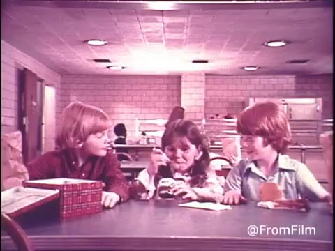 The still captures a nostalgic scene from a 1970s Hunt's Snack Pack Chocolate Pudding commercial. Three kids, two boys and a girl, are seated at a dining table, surrounded by lunchboxes and bright smiles. The girl, in the center, indulges in a Snack Pack pudding, which reflects the playful and carefree spirit of childhood. The background features a simple, retro setting, enhancing the vintage vibe. The tagline emphasizes that Snack Pack makes pudding a party, showcasing the product's fun appeal.