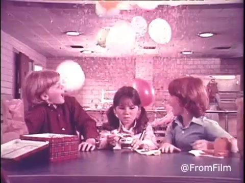 This 16mm still from the 1970s Hunt's Snack Pack Chocolate Pudding commercial captures a playful scene where three children are seated at a table, surrounded by a festive atmosphere. Balloons float above, suggesting a celebration. Two boys, one in a red shirt and another in a light blue, appear excited while interacting with the girl in the middle, who is more focused on her snack. The overall vibe is cheerful and emphasizes the fun of enjoying pudding as part of a party. The nostalgic look reflects the era's style and branding, making it a charming piece of advertising history.