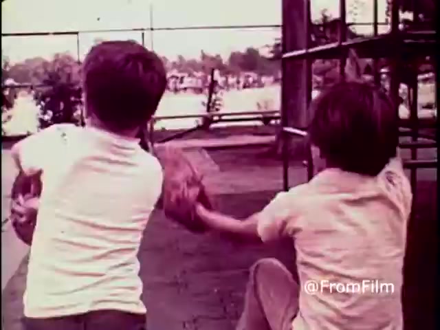 The still from the 1970 PSA 'Why Do You Think They Call It Dope?' depicts two young boys playing with a baseball in a park setting. They are seen from behind, focused on their play, which suggests innocence and the joy of childhood. The warm color tones typical of 16mm prints evoke a sense of nostalgia. The context of the PSA highlights the dangers of drug use, contrasting the carefree nature of the boys with the serious message about substance abuse.