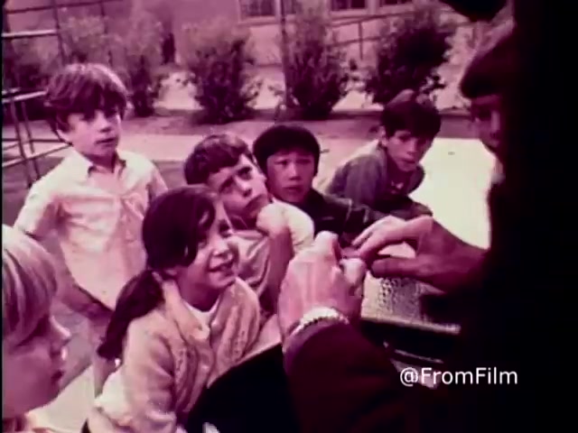 The still from the 1970 PSA 'Why do you think they call it dope?' shows a group of children, intrigued and curious, gathered around an adult who appears to be demonstrating something with their hands. The setting is outdoors, likely in a schoolyard or playground, with sparse greenery in the background. The children's expressions range from amusement to curiosity, highlighting a moment of engagement. The tone of the image suggests a cautionary theme about drugs, typical of anti-drug messaging from that era.