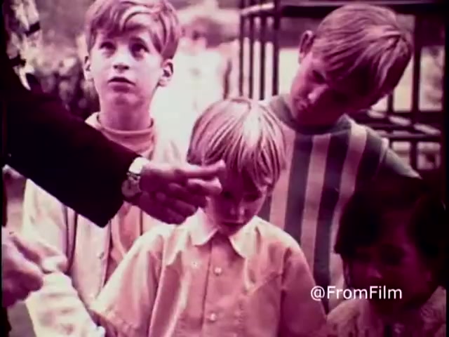 The still from the 1970 PSA 'Why do you think they call it dope?' shows a group of children, with focused expressions, listening intently. An adult’s hand is gesturing towards them, emphasizing a moment of serious dialogue. The muted colors and the children's attire reflect the era, while the scene captures a pivotal moment in the anti-drug message, highlighting the gravity of the conversation. .