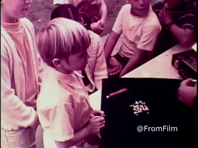 The still from the 1970 PSA 'Why do you think they call it dope?' depicts a scene where a group of children are gathered around a table. A young boy, with light hair and dressed in light clothing, stands focused on a set of pills laid out on a black surface. The adults nearby seem to be explaining the dangers of drugs, capturing the serious tone of the anti-drug message. The visual style characteristic of the era conveys an educational yet impactful atmosphere, aimed at raising awareness among viewers.   .