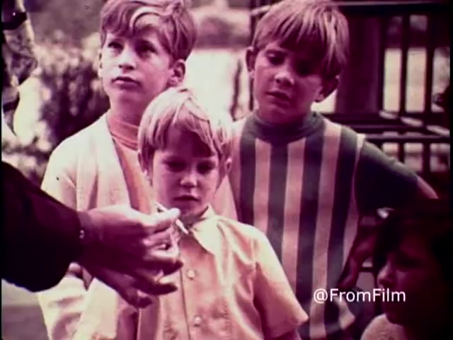 The still captures a tense moment from the 1970 anti-drug PSA 'Why Do You Think They Call It Dope?' featuring a group of young children. They appear concerned or curious as an adult's hand, presumably offering something, is prominently placed in the foreground. The children's expressions convey a mix of intrigue and apprehension, highlighting the serious theme of the campaign. The vintage 16mm print adds a nostalgic quality to the scene, emphasizing the era's approach to discussing drug awareness.   .