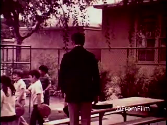 In this still from the 1970 PSA 'Why Do You Think They Call It Dope?', a somber figure in a black coat stands with their back to the camera, overseeing a playground scene filled with children playing. The muted, warm tones of the film enhance the nostalgic yet serious atmosphere, emphasizing the message against drug use. The surrounding environment, marked by chain-link fencing and trees, suggests a school setting, reinforcing the PSA's focus on youth and the impact of drugs.