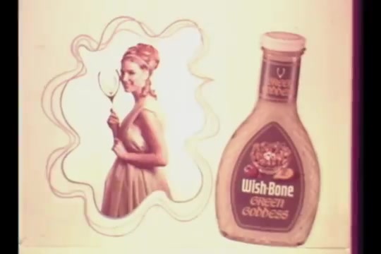 The still features a retro design with a woman in a light-colored dress holding a glass, set against a whimsical outline. To her right, there is a prominently displayed bottle of Wish-Bone Green Goddess salad dressing, showcasing its label and branding. The overall aesthetic reflects the 1960s advertising style, emphasizing freshness and appeal.