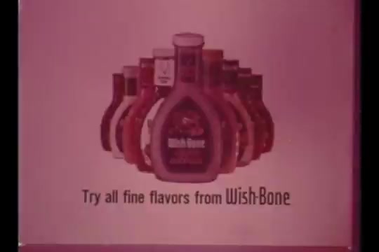 The still features an array of Wish-Bone salad dressing bottles in the foreground, prominently showcasing the Green Goddess dressing. The background has a soft pink hue, and text invites viewers to 'Try all fine flavors from Wish-Bone,' emphasizing the brand's variety. The aesthetic reflects 1960s advertising style, with a focus on enticing consumers.