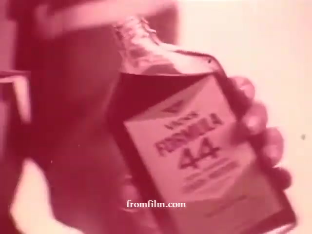 The still features a close-up of a hand holding a bottle labeled 'Formula 44.' The background is blurred, creating a vintage aesthetic typical of 16mm film. The focus is on the product, emphasizing its branding within the context of a commercial.