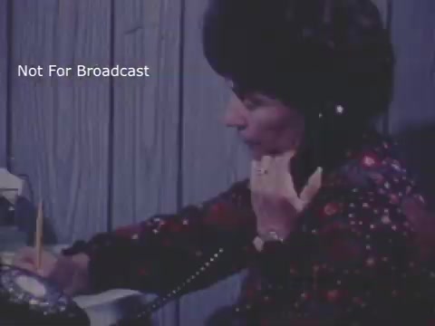 The still depicts a woman in a vintage setting, sitting at a desk and speaking on a telephone. She appears engaged and serious. The background features wooden paneling, and she is holding a pencil, seemingly taking notes. The overall tone suggests a focus on communication and concern, fitting the context of a child abuse PSA from the 1970s. The text 'Not For Broadcast' indicates that this is likely an archival image.