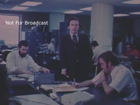 The still shows a 1970s office scene in Wheeling, WV, likely a newsroom or public service setting. A man in a suit stands among people working at desks, engaging with them. The atmosphere appears serious, reflecting the gravity of the child abuse topic addressed in the PSA. The environment has a vintage look, with mid-century office design elements and subdued lighting.