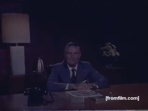 The 16mm still from the 1974 'NYC United Fund Commercial' features a man sitting at a desk in a well-lit office environment. He appears to be professionally dressed, wearing a suit, and is positioned in front of items like a telephone and a stack of books. The backdrop includes a lamp and a small arrangement of flowers, creating a warm, inviting atmosphere. The overall aesthetic reflects the production style of the 1970s, emphasizing a polished, corporate image. This commercial has been reconstructed from separate reels, indicative of the preservation efforts from the former MovieLab film lab.