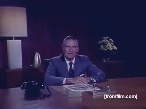 The still from the 1974 'NYC United Fund Commercial' features a man seated at a desk in a formal setting. He is dressed in a suit and tie, with a serious expression, suggesting he is delivering a message of importance. The background includes a lamp and a potted plant, contributing to a professional atmosphere. The desk is organized with some papers, indicating a business context. This footage is part of a reconstructed commercial for what is now known as United Way, emphasizing the organization's approach to community funding during that era.