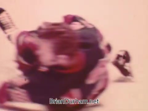 The still features a dynamic hockey scene, showcasing players in action on the ice. One player is sliding, possibly after a collision or play, while another player is in the background. The colors are slightly muted, typical of 16mm film, and the overall composition conveys a sense of movement and excitement associated with hockey.