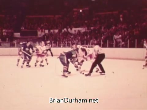 The still captures a moment during an NHL game from 1973, featuring players in jerseys facing off at center ice. A referee is positioned between them, ready to drop the puck. The ice rink is bustling with a crowd in the background, emphasizing the vibrant atmosphere of hockey. The vintage quality of the image reflects the era's broadcasting style.