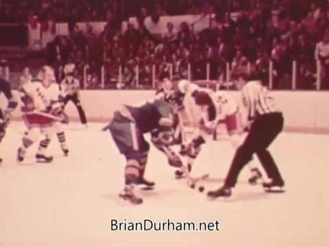 The still captures a moment from a 1973 NHL game, featuring players in traditional hockey gear facing off on the ice. One player in a blue jersey prepares for the puck drop against an opponent in a white jersey. The arena is filled with spectators, creating an energetic atmosphere typical of ice hockey matches. The image reflects the sport's historical style and presentation.