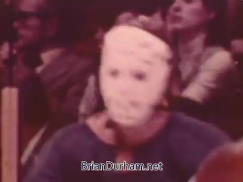 The still features a hockey player wearing a distinctive white mask, typical of the era. The setting appears to be a crowded arena, likely during a game, with spectators visible in the background. The image captures the intense atmosphere of hockey in the 1970s.