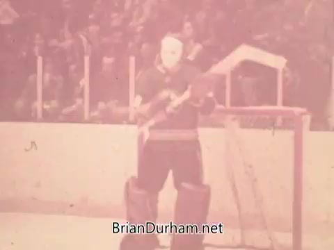 The still features a goalie in a hockey rink, wearing a mask and hockey gear. The scene is set against a backdrop of a packed audience in the stands. The atmosphere suggests a moment of anticipation, likely before a game or during a break, typical of promotional content from the 1973 NBC NHL Bumper Promo. The color is faded, characteristic of 16mm film from that era.