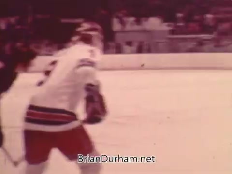 The still features a hockey player in a white jersey with blue and red accents, indicating a team uniform, likely from the NHL. The player appears focused on the game, with an opponent nearby. The ice rink setting suggests an action-packed scene typical of promotional content. The background features some spectators, hinting at a lively atmosphere during a match.