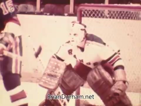 The still features a goalie in vintage hockey gear, crouched in a ready stance on the ice. He is wearing a white jersey with multi-colored stripes, and the scene captures a moment during a hockey game, likely emphasizing the tension and anticipation typical in sports. The background shows blurred outlines of spectators and the rink, adding to the nostalgic feel of the 1973 NHL promo.