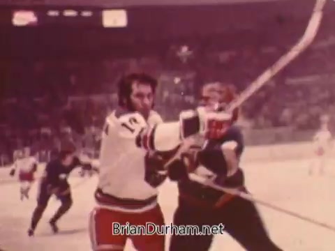 The still from the '1973 NBC NHL Bumper Promo' depicts an intense moment during a hockey game. In the foreground, two players engage in a physical confrontation, with one player in a white jersey holding a hockey stick. The background shows the hockey rink and other players moving, emphasizing the action-packed nature of the sport. The colors are slightly faded, typical of 16mm film from that era.