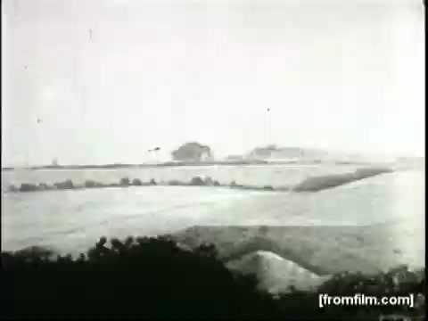 The still from the 'Movietone Battle Of Britain' newsreel features a panoramic view of a landscape, showcasing rolling hills and fields. The scene is rendered in black and white, typical of the era, capturing a wartime atmosphere. The image likely features aircraft in the distance, representative of the battle context, while the surrounding terrain suggests a rural setting in Britain during World War II.