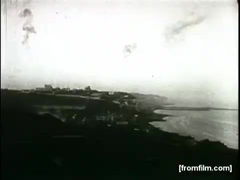 The still from the 'Movietone Battle of Britain' newsreel features a coastal scene, likely depicting British land near the water. The image captures a somewhat foggy or overcast atmosphere, with various structures visible along the coastline. The quality is characteristic of 16mm film, showing grain and a lack of sharpness. This frame serves as a historical representation of the setting during the Battle of Britain, narrated by Lowell Thomas.