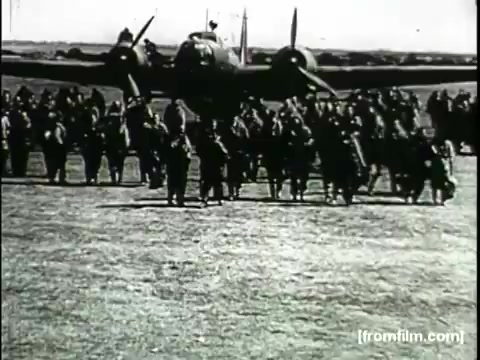 The still depicts a significant moment from the 'Movietone Battle of Britain' newsreel. It shows a large group of soldiers gathered on an airfield, with a military aircraft in the background. The composition emphasizes the presence of the troops, likely symbolizing solidarity and readiness in the context of the Battle of Britain. The black-and-white print suggests a historical perspective on wartime efforts, often associated with the reporting style of Lowell Thomas.