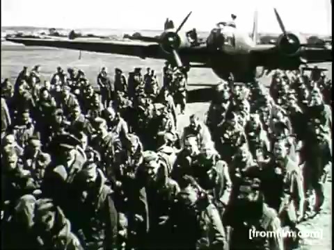 The still from the 'Movietone Battle of Britain' newsreel depicts a large group of soldiers gathered in a field, some appearing to salute or cheer. In the background, a military aircraft is visible, suggesting a wartime setting. The image conveys a sense of camaraderie and determination among the troops during a significant moment in history.