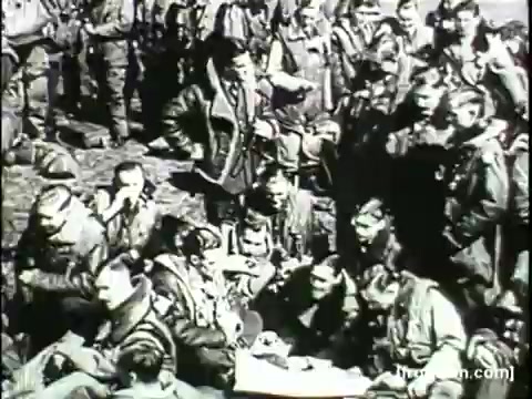 The still from the 'Movietone Battle of Britain' newsreel features a group of military personnel, likely pilots, gathered together in a field. They are dressed in flight jackets and appear to be engaged in conversation or preparing for their next mission. The atmosphere conveys camaraderie amidst the backdrop of the wartime context, capturing a moment of solidarity among the soldiers.