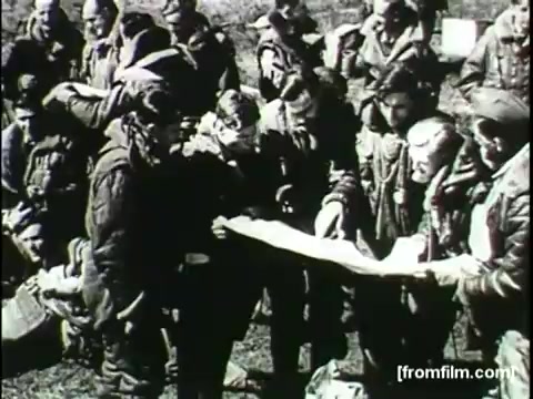 The still from the 'Movietone Battle Of Britain' newsreel shows a group of military personnel gathered around a map. They appear engaged in discussion, likely strategizing or planning. The scene captures the intensity of wartime decision-making, with the soldiers dressed in flight gear and displaying expressions of focus and determination. The black-and-white aesthetic of the 16mm print adds a historical and documentary feel to the image.