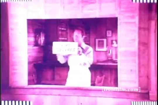 The still features Bernie Kopell in a rustic, wooden setting typical of beer commercials from the early 70s. He holds a sign that likely promotes Old Milwaukee Beer, enhancing the lighthearted and jovial atmosphere of the ad. The color tones are warm and nostalgic, characteristic of the era's advertisements.