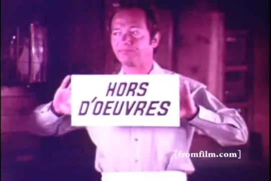 The still features a man, likely Bernie Kopell, presenting a sign that reads 'HORS D'ŒUVRES.' He is dressed in a light-colored shirt, holding the sign with a serious expression. The background appears rustic, possibly resembling a cabin or pub setting, and the overall color tone gives off a warm vibe, typical of vintage commercials from the early 1970s.