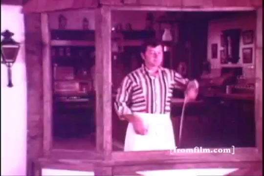 The still from the 1971 Old Milwaukee Beer commercial features a man in a striped shirt and white apron, standing behind a rustic wooden bar. He appears to be serving or preparing something, likely beer, in a lively setting filled with old-fashioned decor. The overall tone is warm and inviting, capturing a classic pub atmosphere.