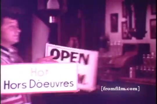 The still from the 1971 'Old Milwaukee Beer' commercial features a man holding two signs: one displaying 'OPEN' and the other reading 'Hot Hors D'oeuvres.' The setting appears to be a cozy bar or tavern. The vibrant colors typical of 16mm film enhance the nostalgic feel of the advertisement. Bernie Kopell's involvement adds a recognizable presence to the piece, which likely aims to convey a warm, inviting atmosphere alongside fine beer and snacks.