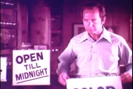The still from the 1971 'Old Milwaukee Beer' commercial features Bernie Kopell in a casual setting. He is shown holding a sign that reads 'COLOR,' with another sign stating 'OPEN TILL MIDNIGHT' prominently displayed nearby. The atmosphere appears warm and inviting, characteristic of vintage commercials from that era. The color tone suggests a nostalgic feel, typical of 16mm prints.
