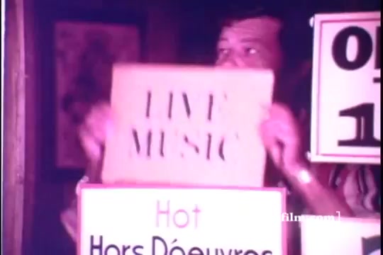 The still captures a moment from the 1971 Old Milwaukee Beer commercial featuring Bernie Kopell. In the scene, a man is prominently holding a sign that reads 'LIVE MUSIC,' while surrounded by various signs advertising food. The atmosphere suggests a lively venue, highlighting entertainment and hospitality, characteristic of beer advertisements of that era.