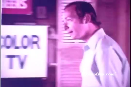 The still shows Bernie Kopell, likely in a light-colored shirt, engaging in the scene of an Old Milwaukee Beer commercial from 1971. Behind him is a sign that reads 'COLOR TV,' suggesting the era's style and setting. The image captures a lighthearted moment, characteristic of vintage advertisements, conveying a sense of warmth and camaraderie associated with enjoying a beer. The coloration hints at the film's age, typical of 16mm prints.