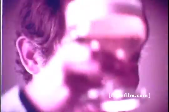 The still features a blurred close-up of a man holding a glass of beer, likely toasting or gesturing to the camera. The lighting has a soft, warm hue, creating a nostalgic atmosphere. The focus is primarily on the glass, emphasizing the beverage, while the man's face remains slightly obscured. The overall composition captures the charm typical of 1970s commercials.