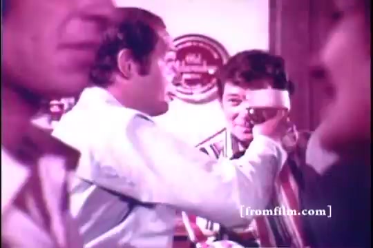 The still from the 1971 Old Milwaukee Beer commercial features Bernie Kopell engaging with another man in a lively bar setting. Kopell holds a beer glass, smiling and appearing to toast or share a moment with the other character. The background is filled with indistinct figures, creating a friendly, social atmosphere characteristic of beer advertisements from that era.