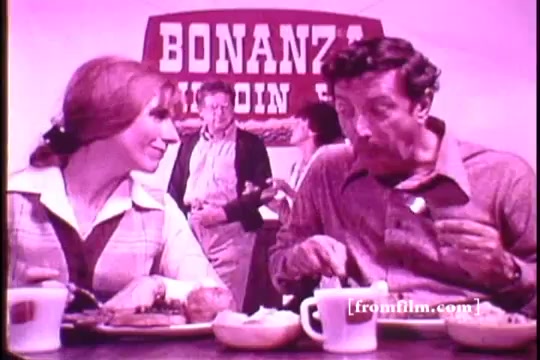 The still from the Bonanza Restaurant commercial features a woman and a man sitting at a table, enjoying meals. The man is eating, while the woman smiles at him. In the background, another figure appears, and a large sign reading 'BONANZA' is prominently displayed. The visual style reflects the 1960s era, likely with a warm color palette. The setting emphasizes the casual dining atmosphere of the Bonanza Steakhouse chain, which was founded by Dan Blocker in 1963.